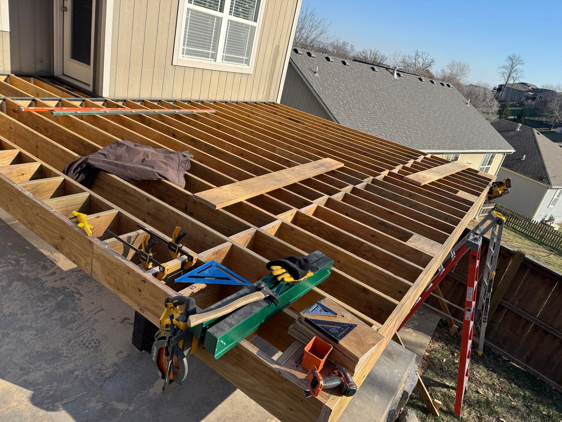 Roof deck framing under construction beside a house, with lumber, tools, and a ladder.