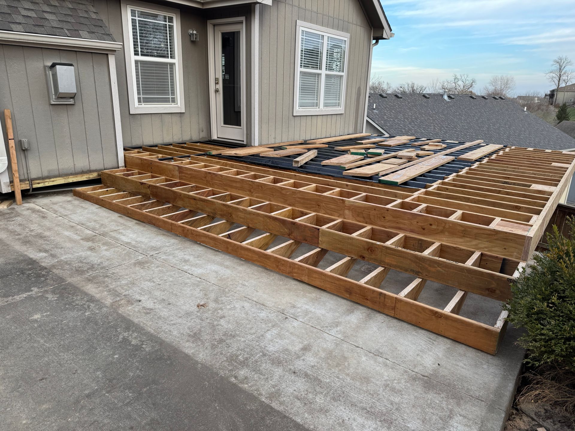Partially built wooden deck frame beside a house, with concrete driveway in the foreground.