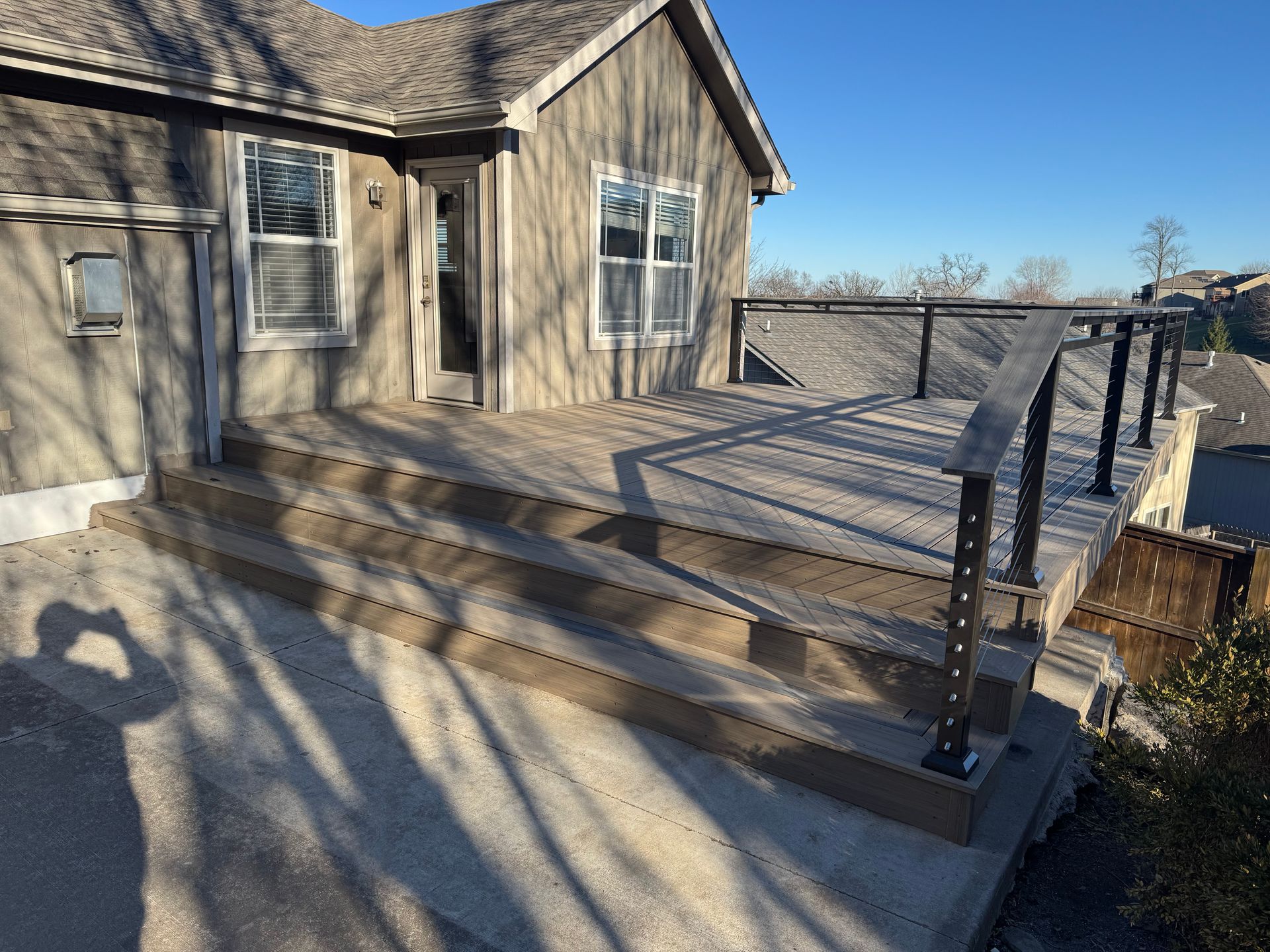 House with steps and wooden deck beside a driveway, with long tree shadows in bright daylight