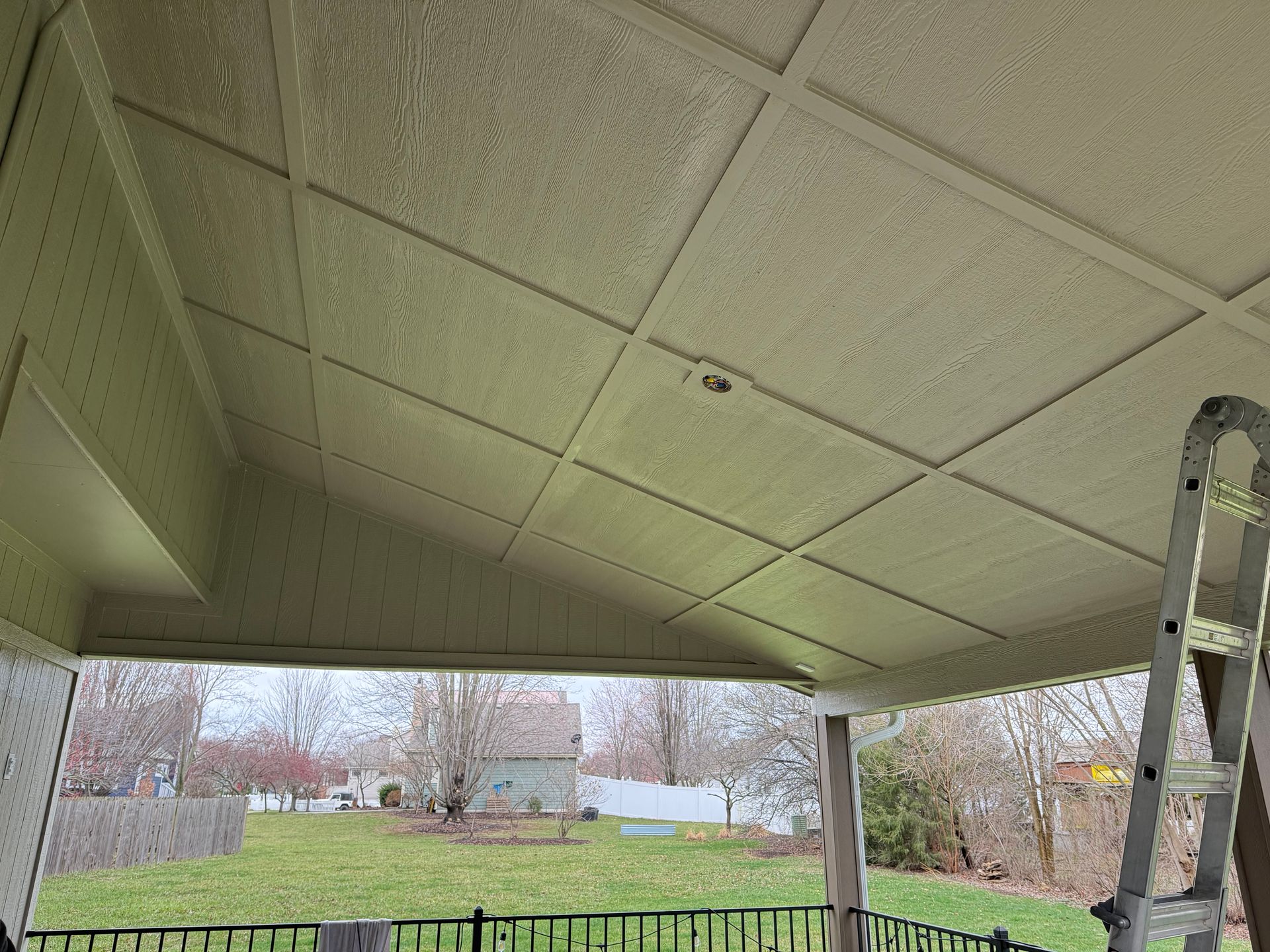Covered porch ceiling with a ladder at right, overlooking a grassy yard and trees beyond.