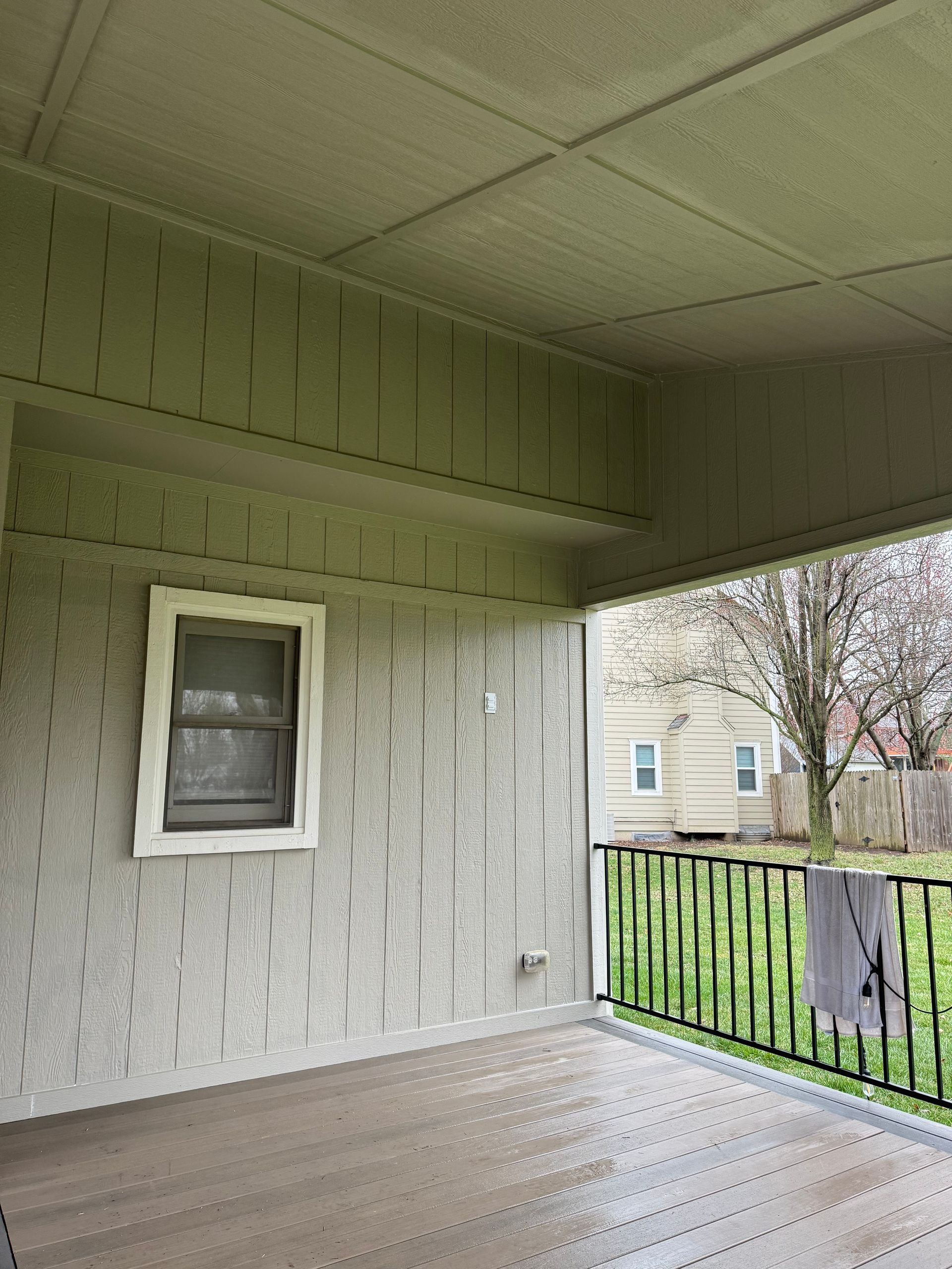 Covered porch with gray siding, a small window, and a black railing overlooking a yard and neighboring house.
