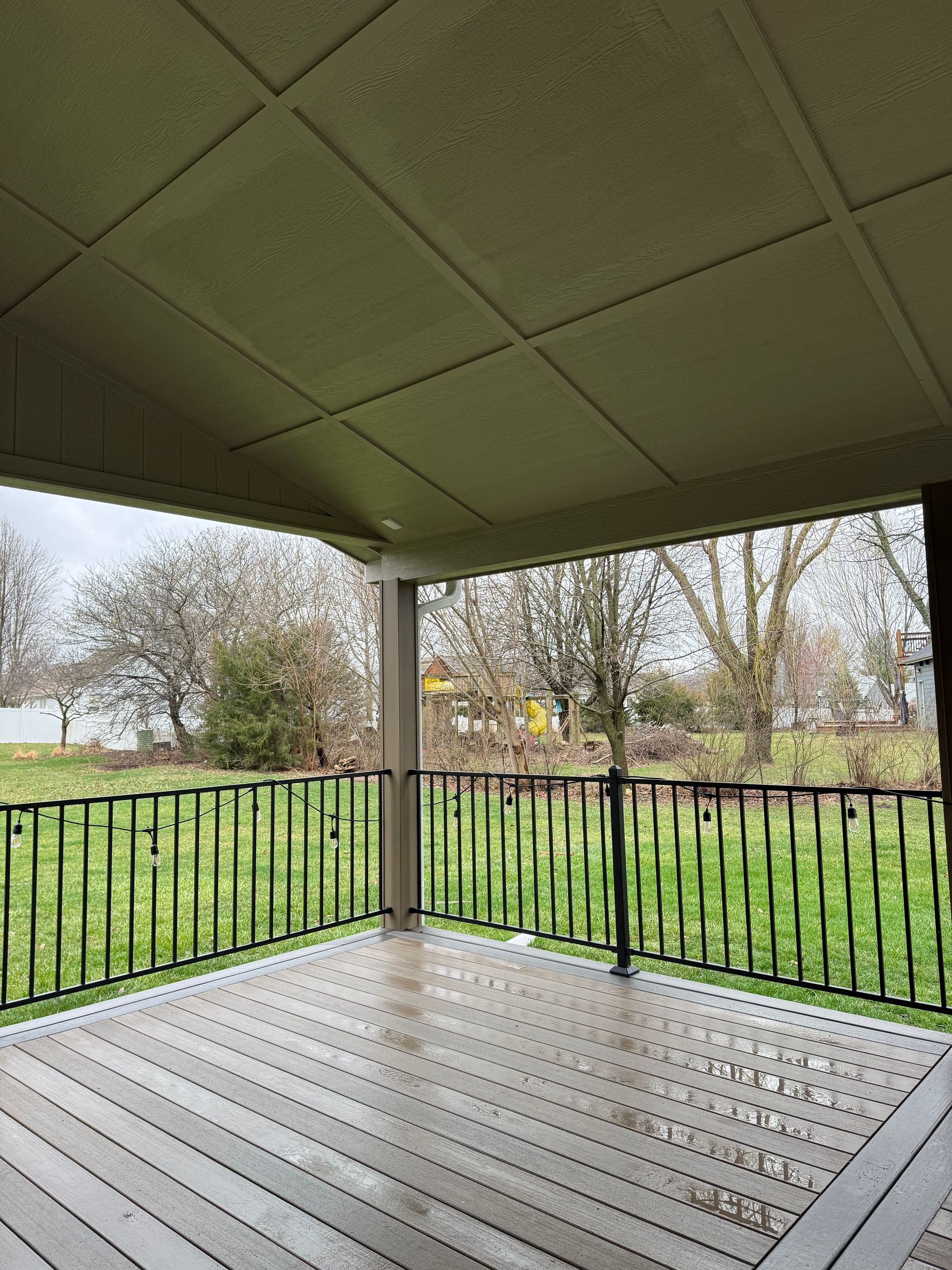 Covered porch overlooking a green yard with black railing and wet gray floor, trees in the background