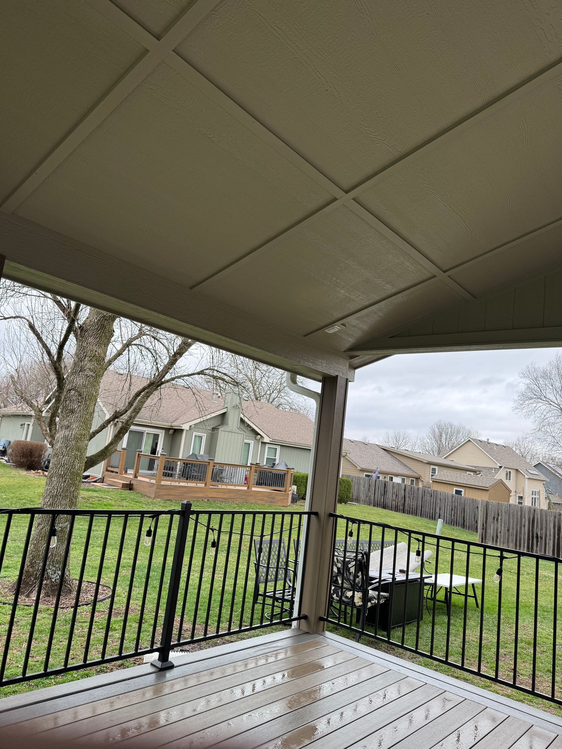 Covered backyard porch with black railing overlooking a grassy yard and neighboring houses.