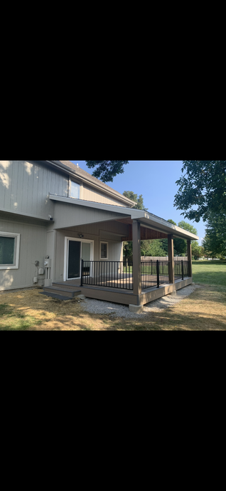 Gray house with a covered porch and wooden railing, shaded by trees on a sunny day