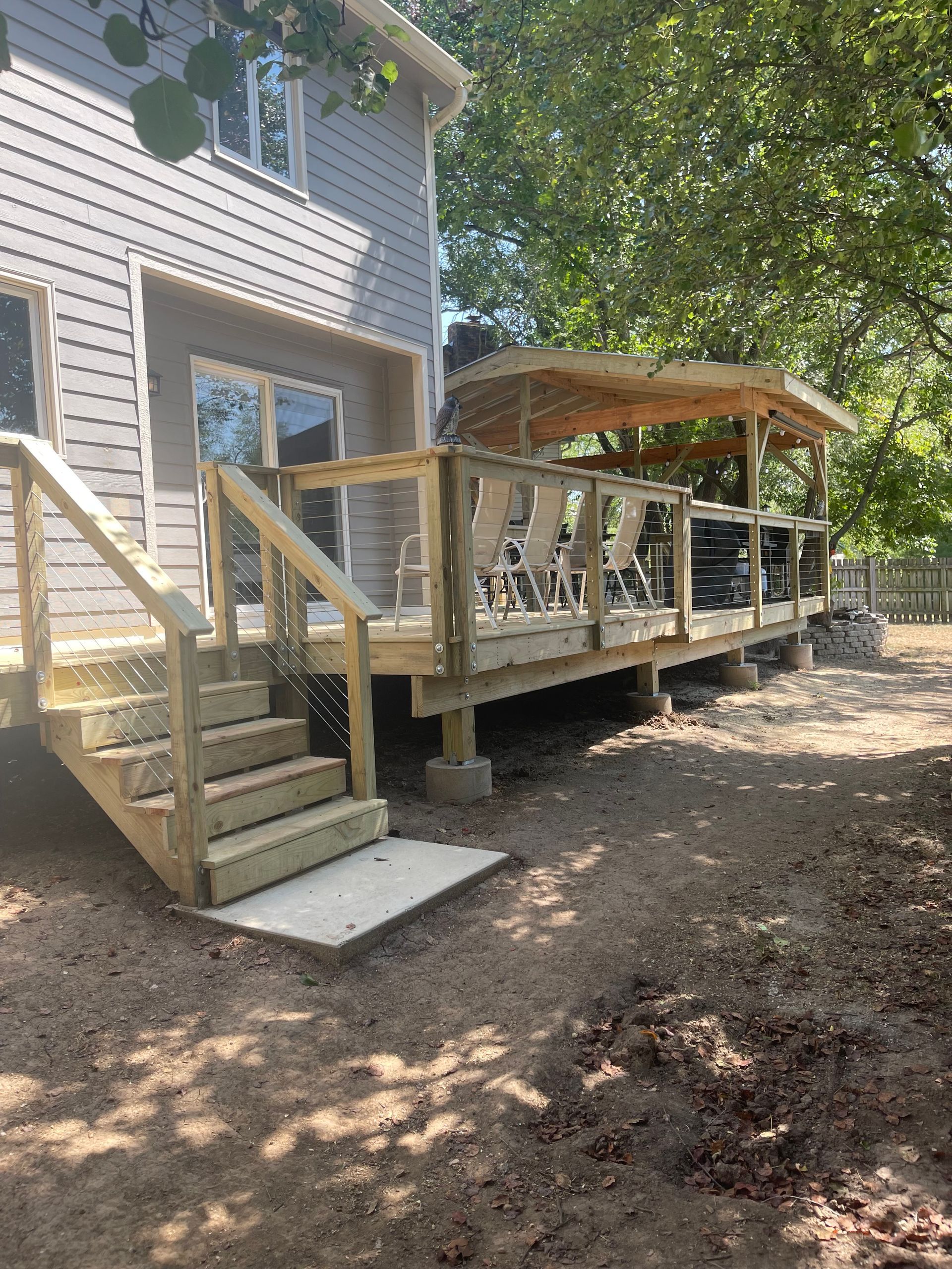 Wooden deck with stairs and ramp beside a house under trees, with a covered seating area.