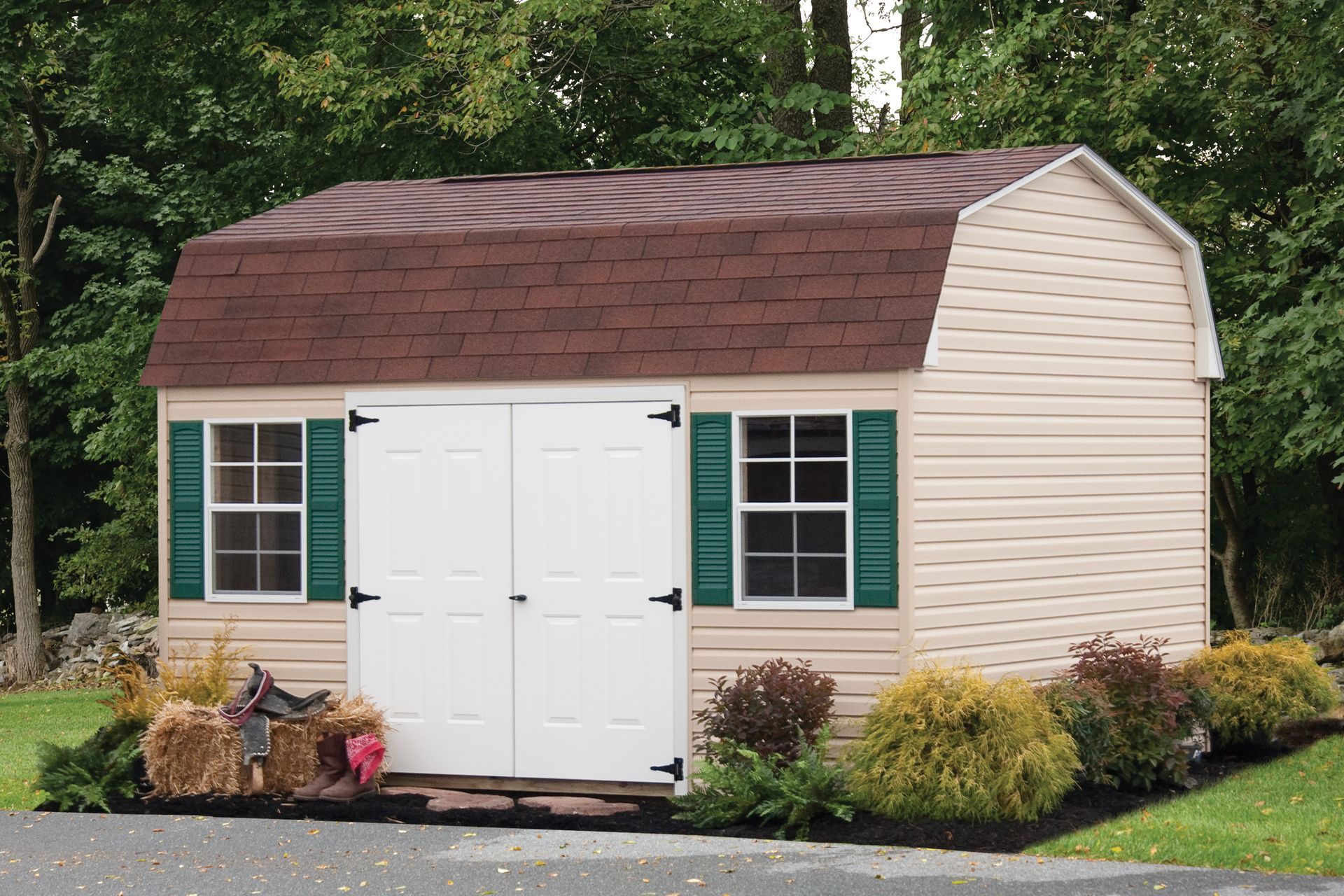 Beige garden shed with white double doors, green shutters, and a brown roof in a landscaped yard