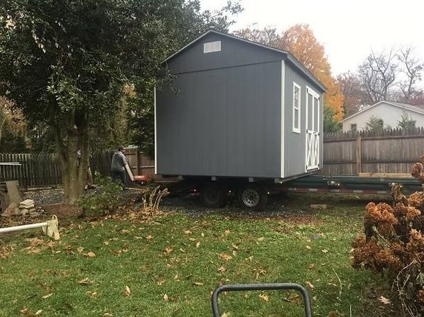 Gray tiny house on wheels in a backyard, with autumn trees and a fence in the background.