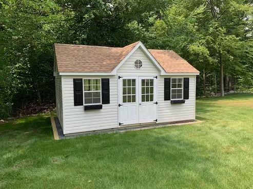 Small white backyard shed with black shutters and double doors on a grassy lawn by trees