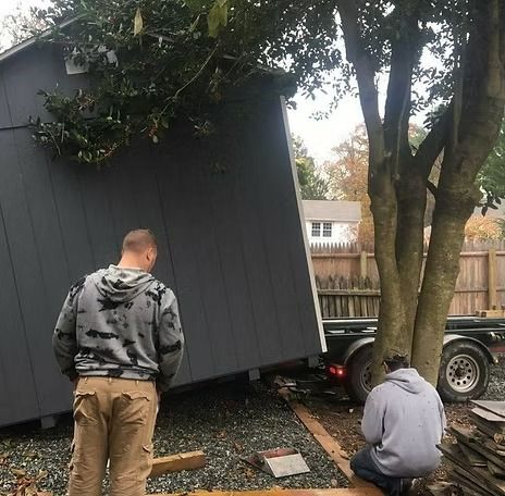 Two men inspect a tilted gray shed near a tree and trailer in a yard