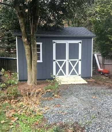Gray garden shed with white double doors and a ramp, beside a tree and gravel path.