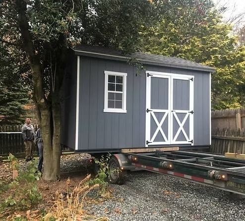 Gray shed with white trim being moved on a trailer in a wooded yard