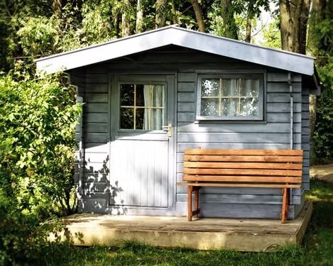 Small gray garden shed with white door and window, front bench, surrounded by trees and greenery.