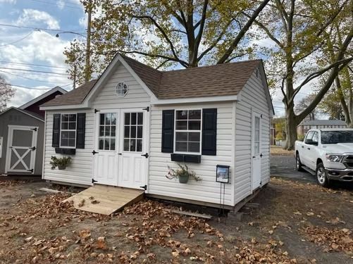 Small white cottage with black shutters, front door, and ramps, in a leaf-covered yard with a van nearby