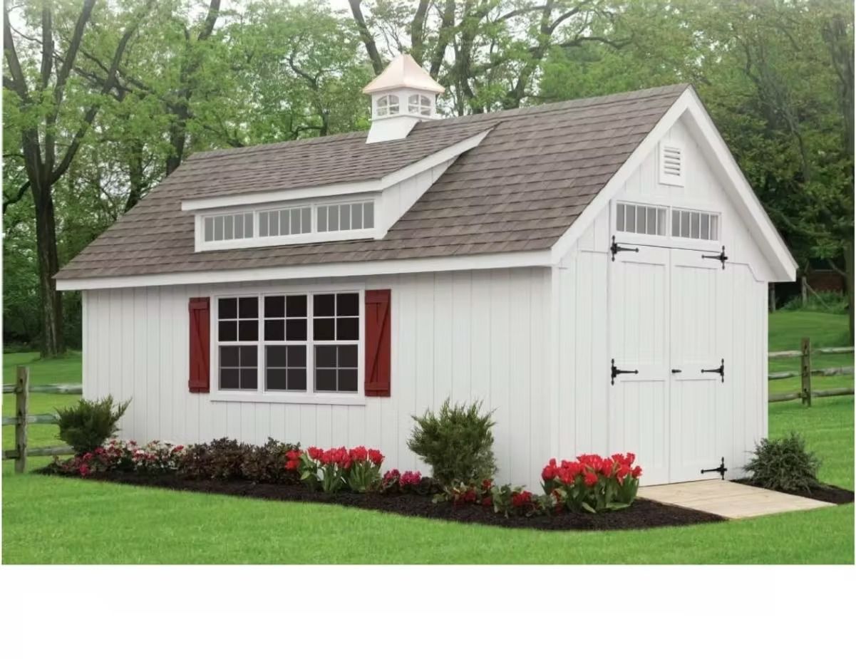 White garden shed with red shutters and flower beds in a grassy yard, surrounded by trees.