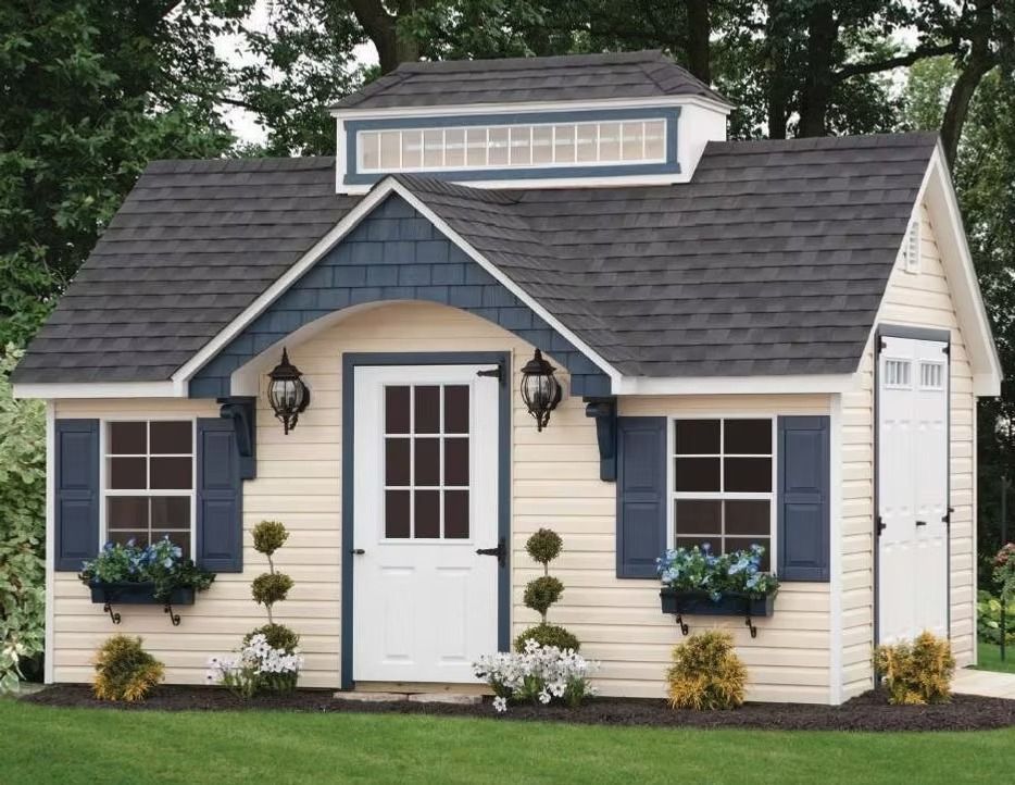 Small cream house with dark roof, blue shutters, white door, and flower planters on the front lawn