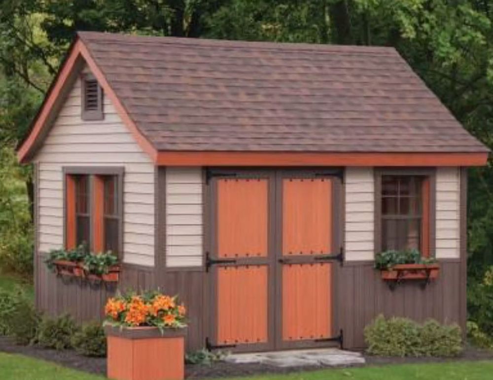 Small gray garden shed with orange double doors, flower boxes, and a potted plant in a wooded yard