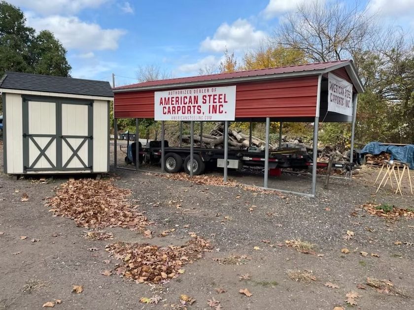 Red steel canopy shelter with “American Steel Carports, Inc.” sign beside a small shed and trailers outdoors