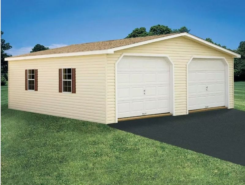Tan beige detached garage with two white doors, small windows, and a shingled roof on a grassy lot.