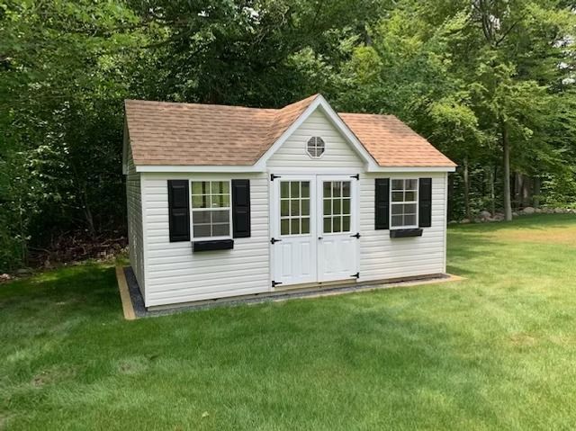 Small white shed with black shutters and tan roof on a green lawn, surrounded by trees