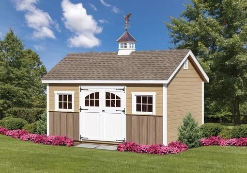 Small beige garden shed with white double doors, pink flower border, and cupola in a grassy yard.