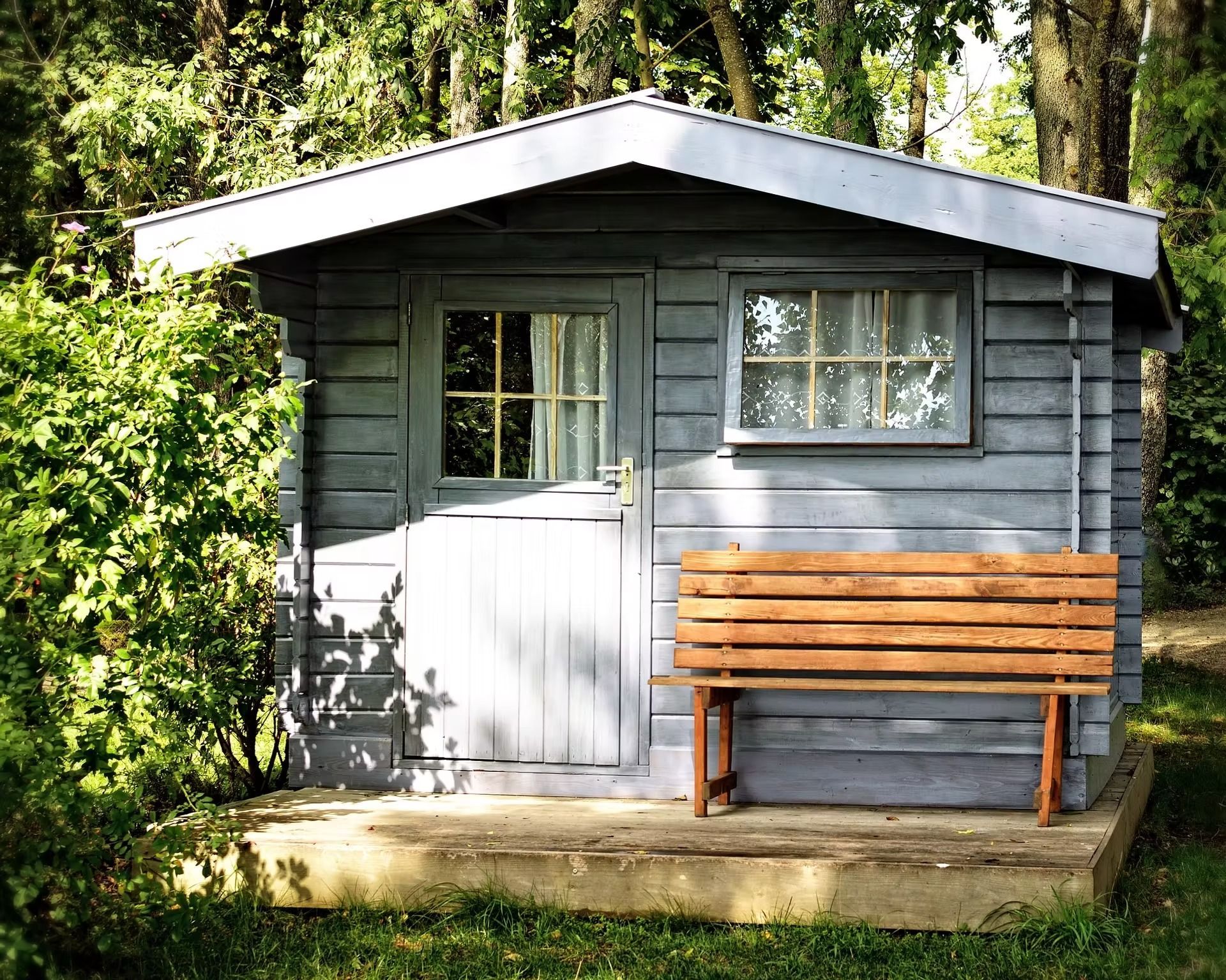 Small gray wooden cabin with a white door and a bench, surrounded by trees and greenery.