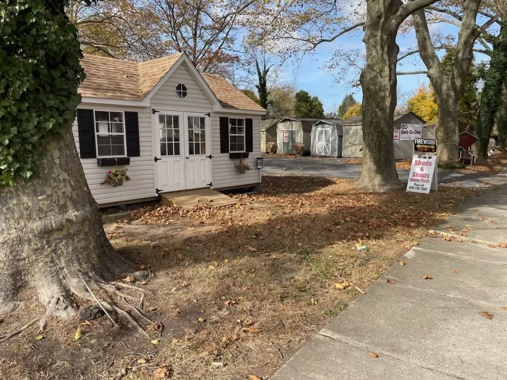 Small white house with black shutters beside a sidewalk, with fall leaves and a yard sign out front.