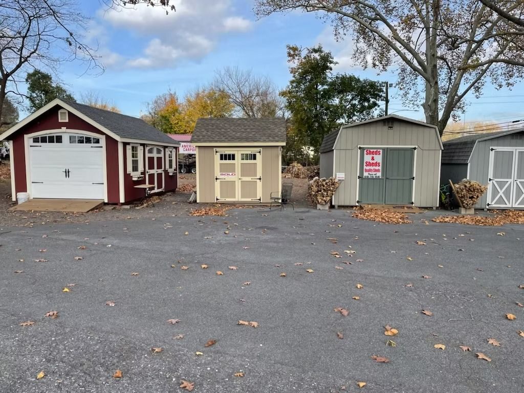 Row of small sheds and a garage in a parking lot, with fall leaves on the ground.