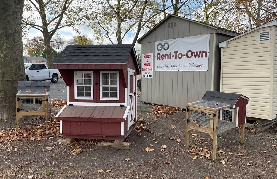 Small red rent-to-own playhouse beside a gray shed and beige building outdoors in fall.