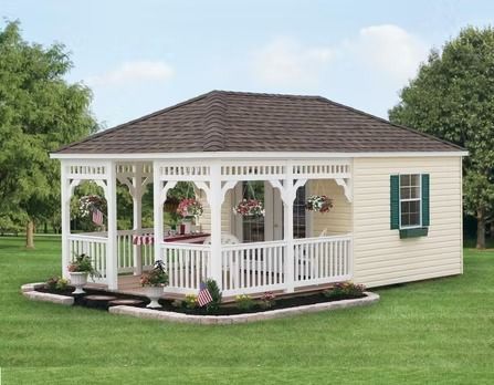 Small beige gazebo-style shed with white trim, green shutters, and a wraparound porch on a lawn.