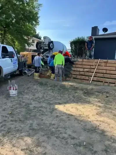 Workers pouring concrete beside a white truck at a construction site, with wooden forms and a mixer truck.