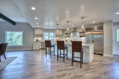Bright modern kitchen and open living area with white island, stools, stainless steel appliances, and hardwood floors