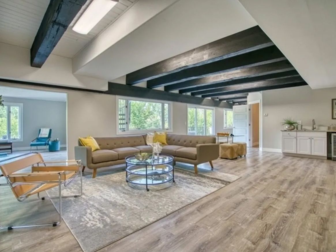 Modern living room with exposed beams, tan sectional, glass coffee table, and bright windows
