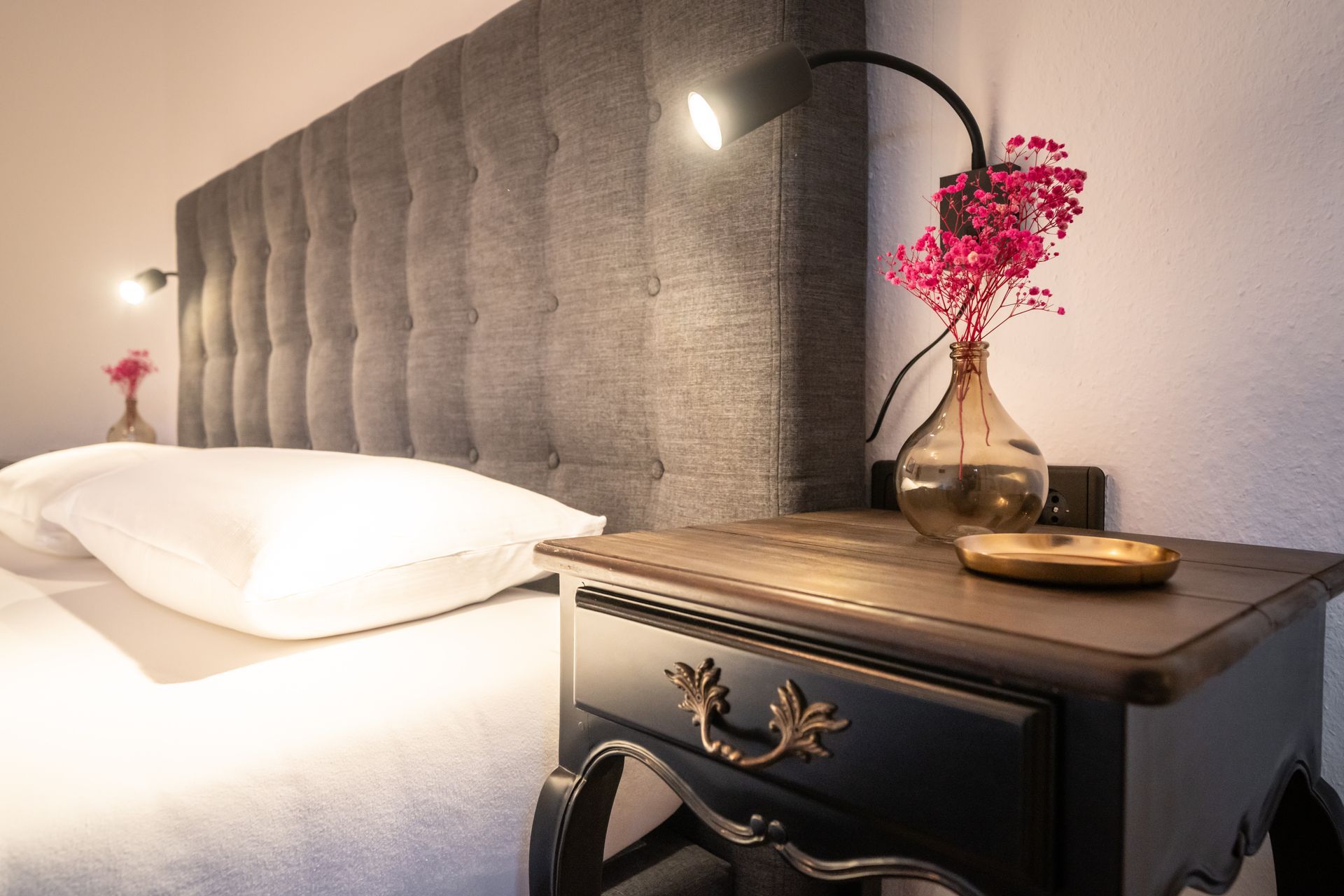 A nightstand with a vase of flowers on it next to a bed.