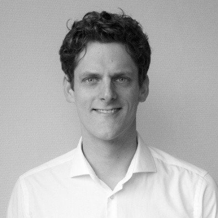 Een man in een wit overhemd lacht naar de camera, tegen een neutrale achtergrond.