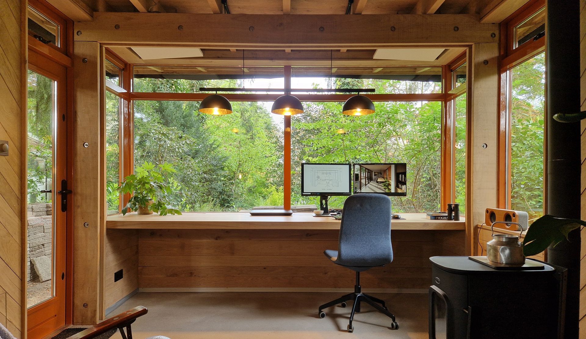 Een comfortabel thuiskantoor met een groot raam dat uitkijkt op weelderig groen. Bureau met computer, stoel en hanglampen.