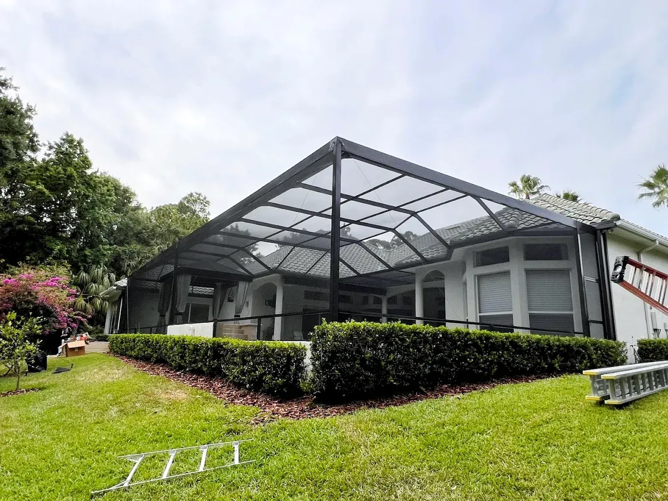 Screened-in patio with black frame, attached to a white house. Green bushes and grass in foreground, trees in the background.