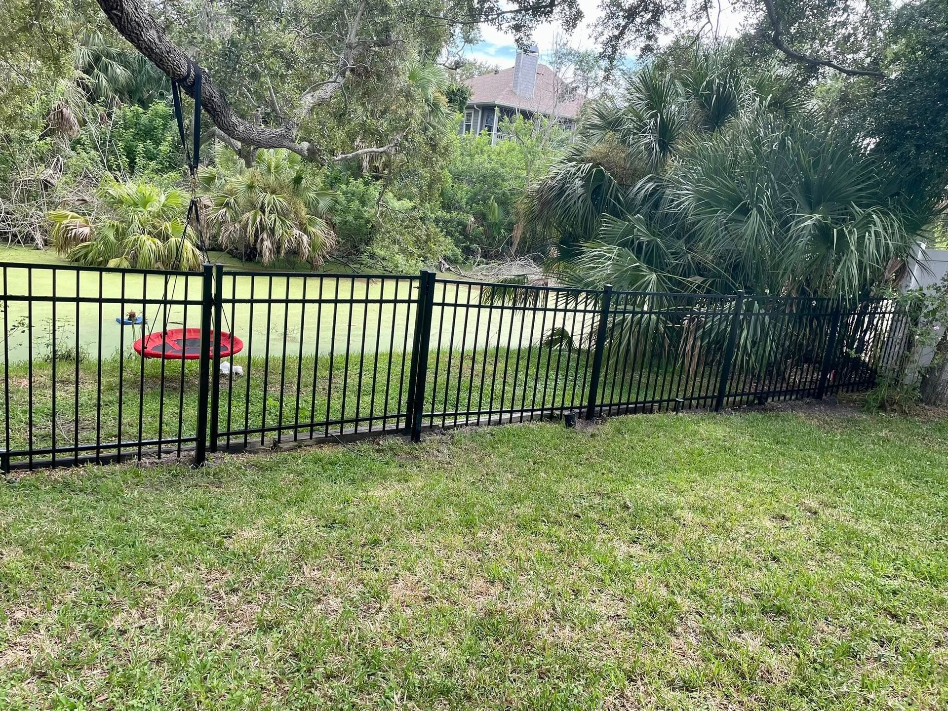 Black metal fence borders a grassy yard, with a pond and lush foliage visible behind it.