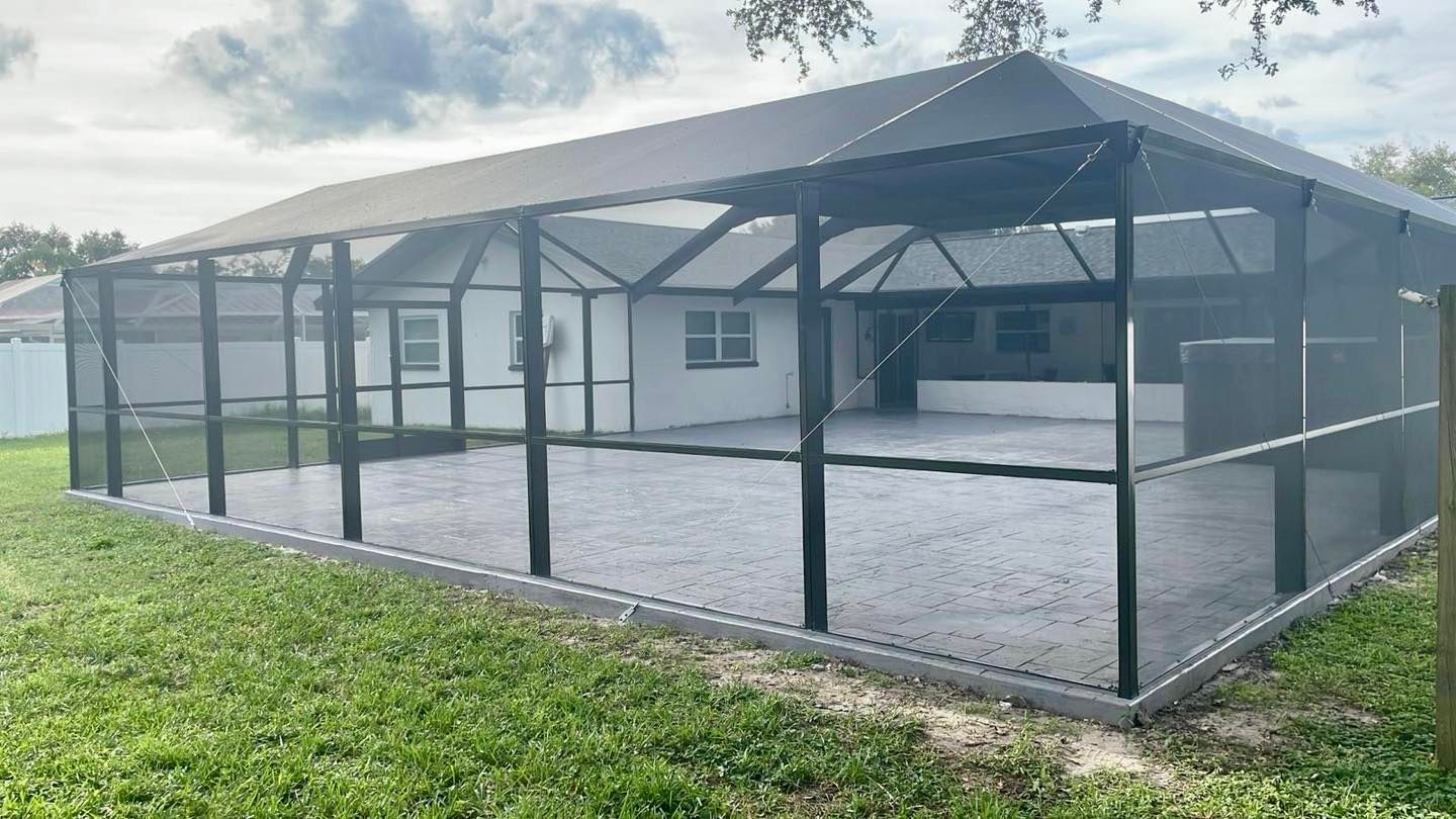 A large, screened-in patio enclosure in a backyard; black frame, gray floor, white house background.