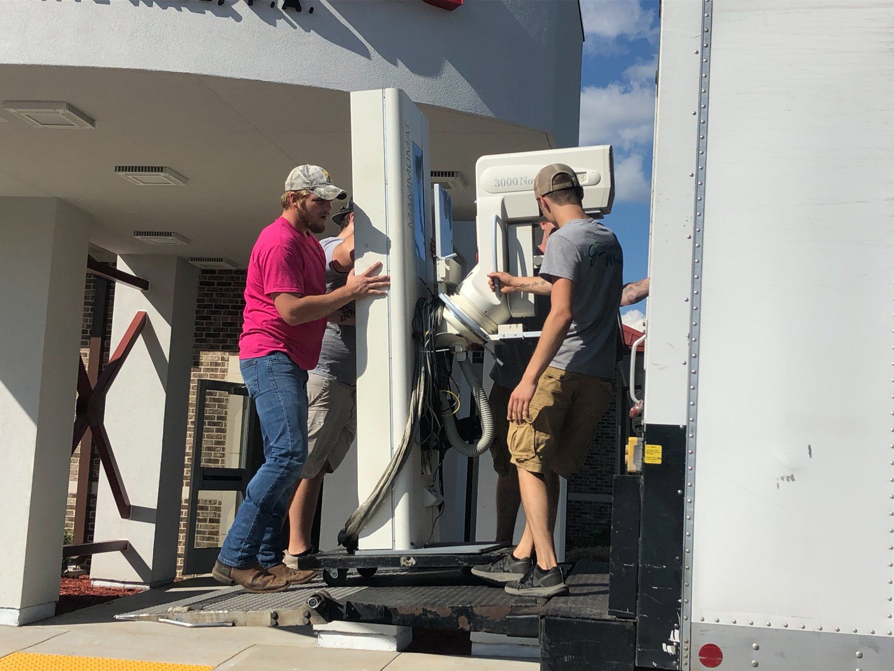 Two workers unload a large machine from a truck at a building entrance.
