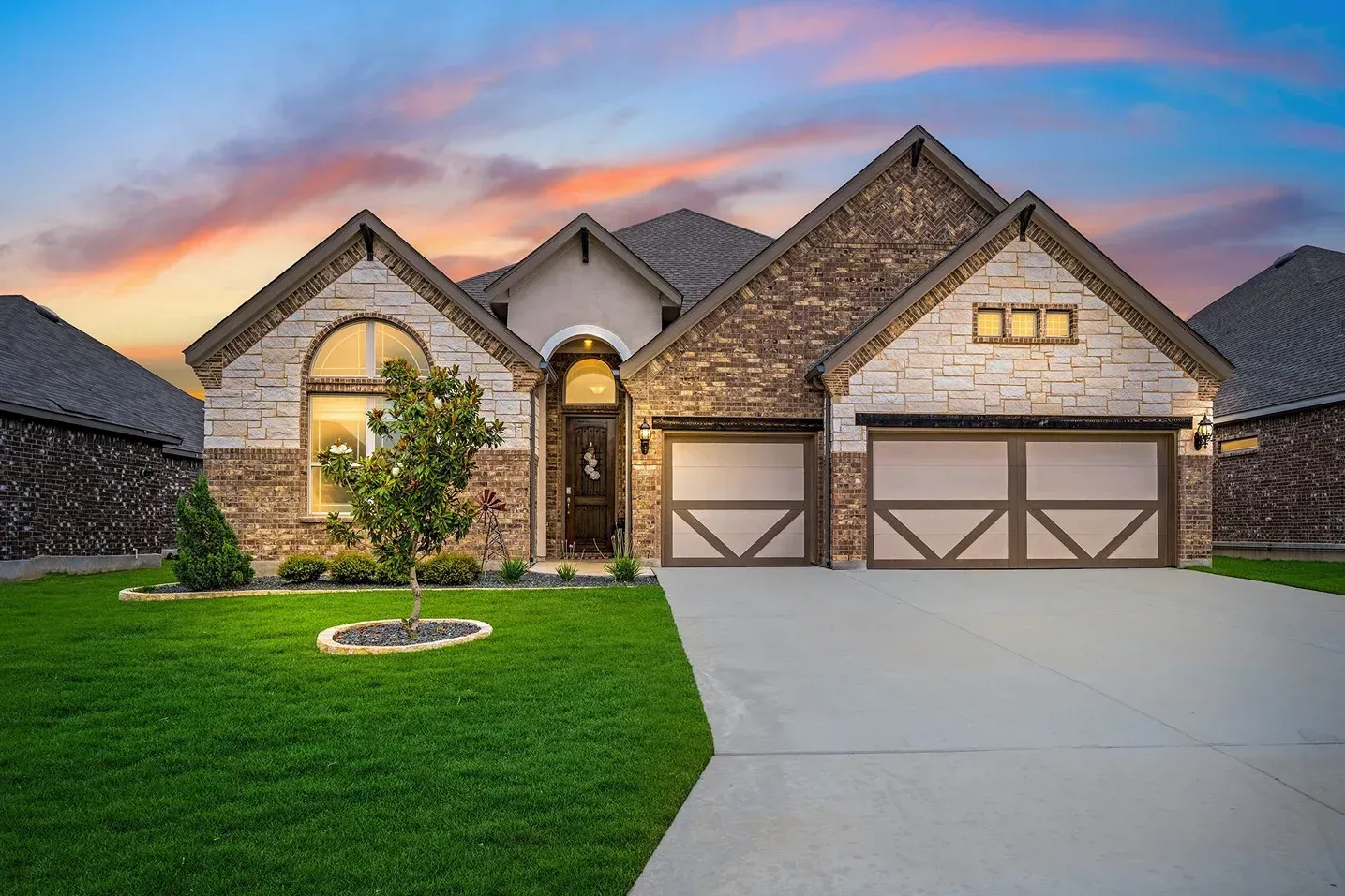 Modern stone suburban house at dusk with a green lawn and two-car garage, warm lights glowing inside