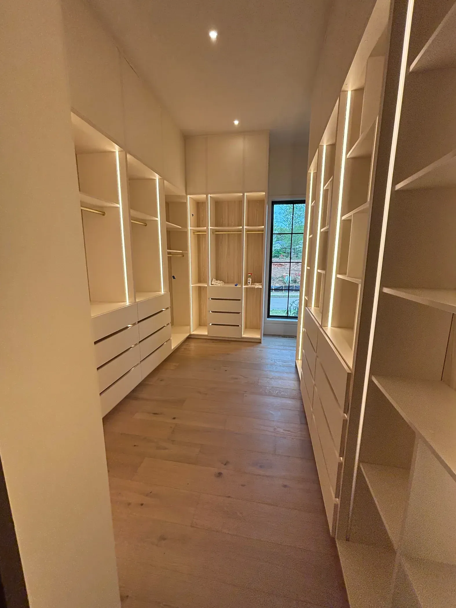 Long, narrow walk-in closet with white built-in shelves and drawers, wood floor, and a window at the end.