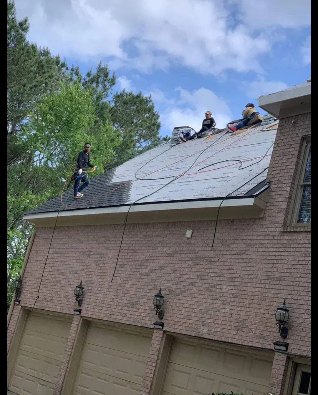 Workers on a steep roof securing shingles with ropes beside a brick garage under a blue sky