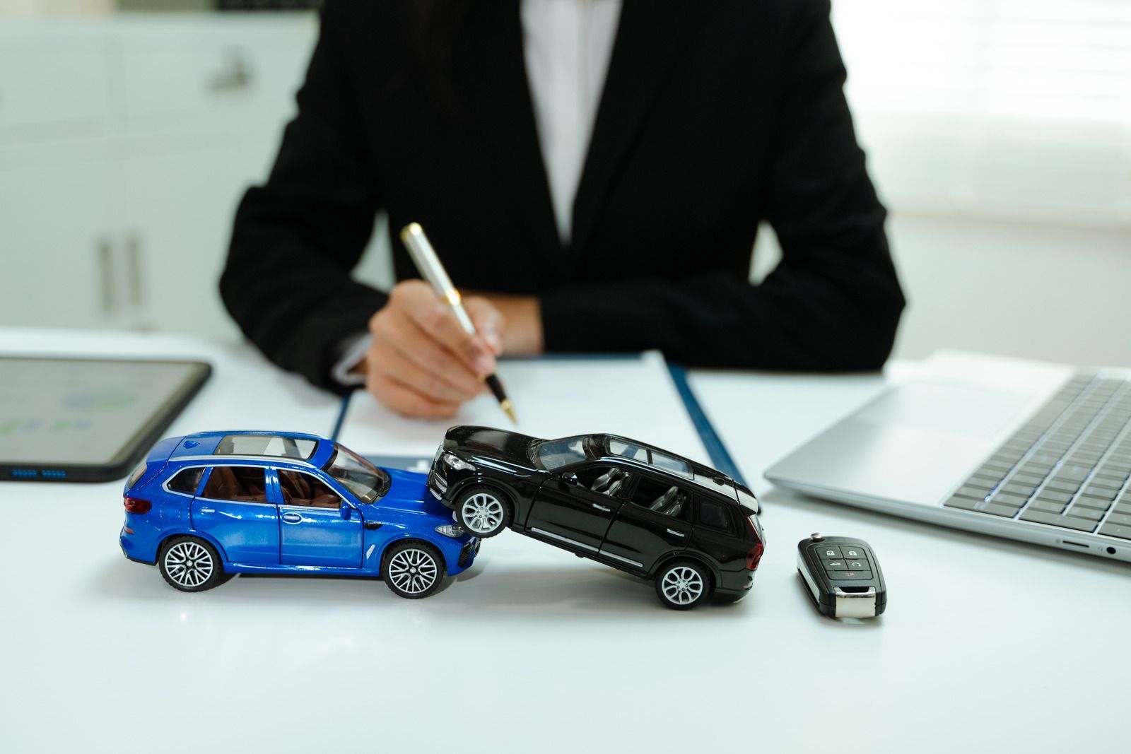 Toy cars on a desk as a person in a suit writes behind them, with a laptop nearby.