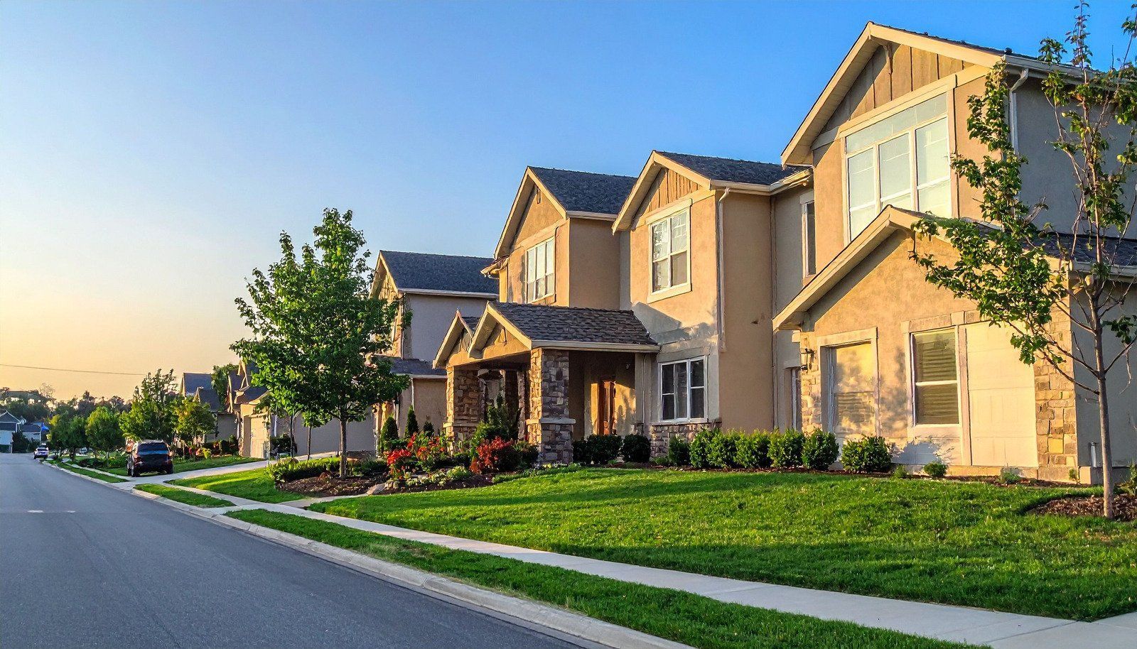 Suburban houses along a quiet street at sunset, with manicured lawns and trees.