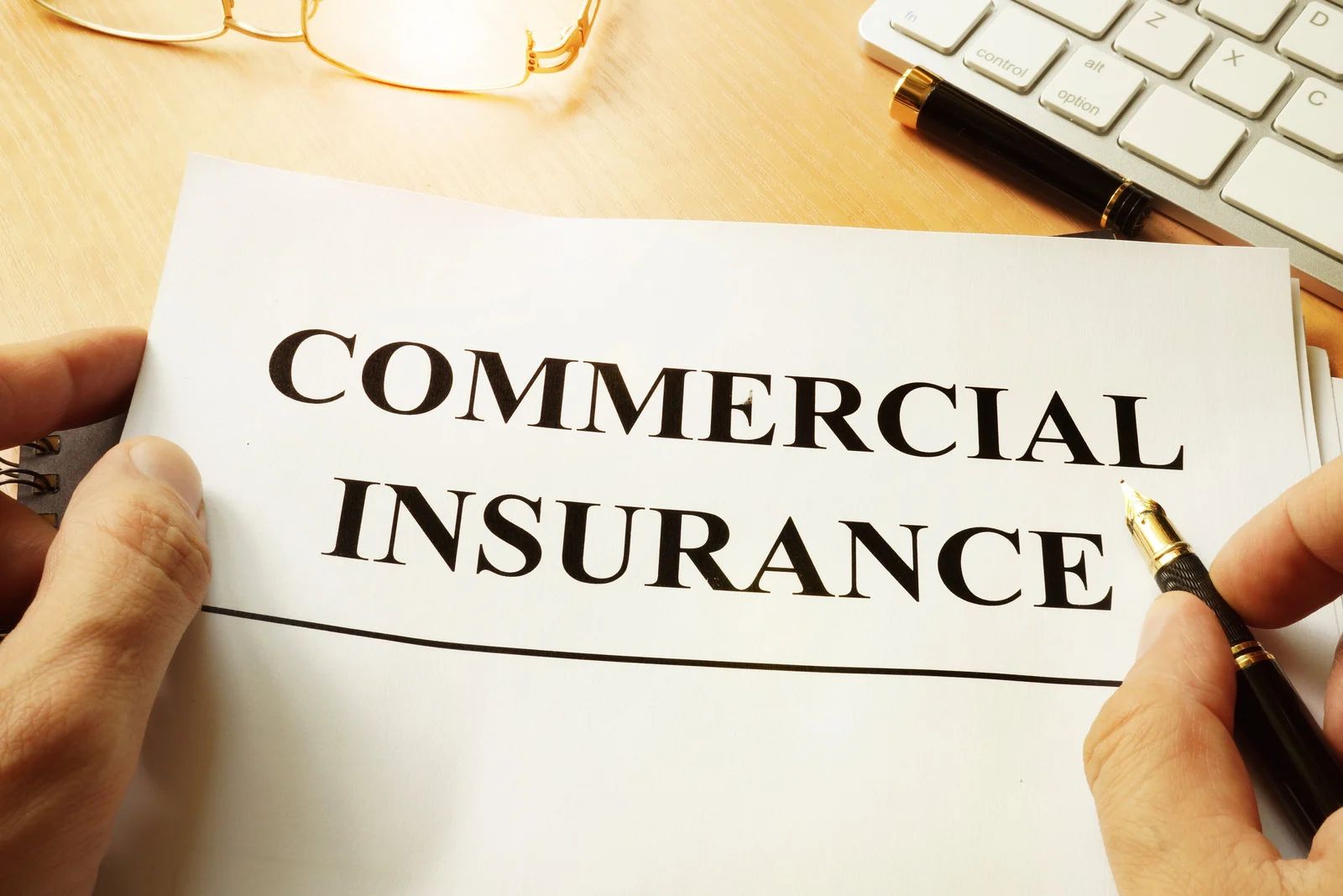 Hands holding a paper labeled “COMMERCIAL INSURANCE” beside a pen on a desk.