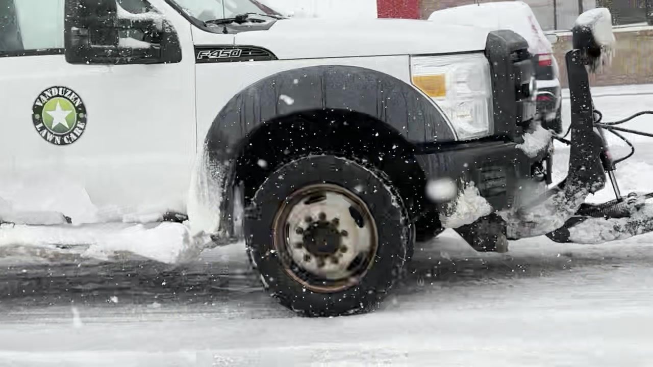 White utility truck with city logo driving through snowy street, front wheel splashing slush