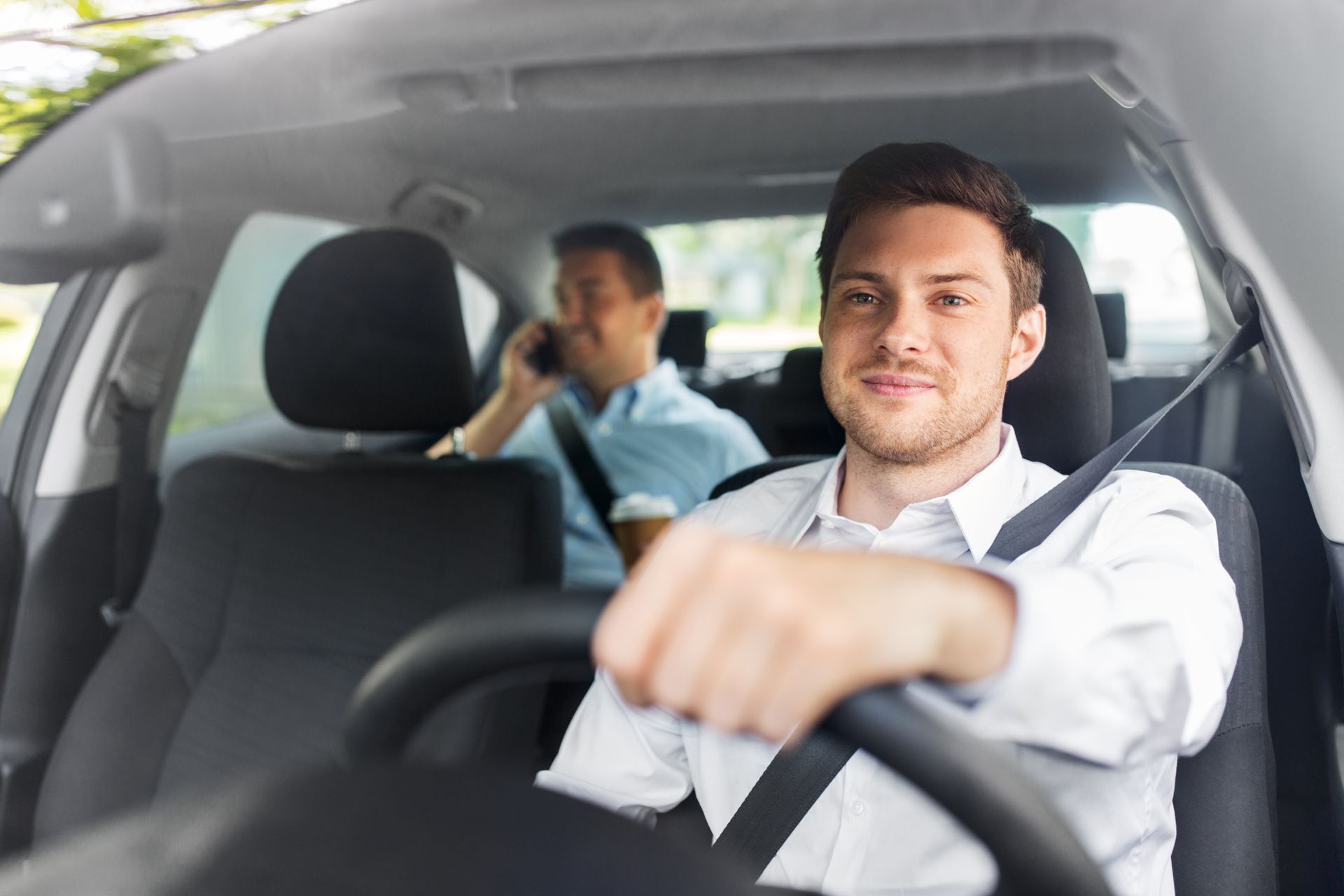 A man is driving a car with another man in the back seat talking on a cell phone.