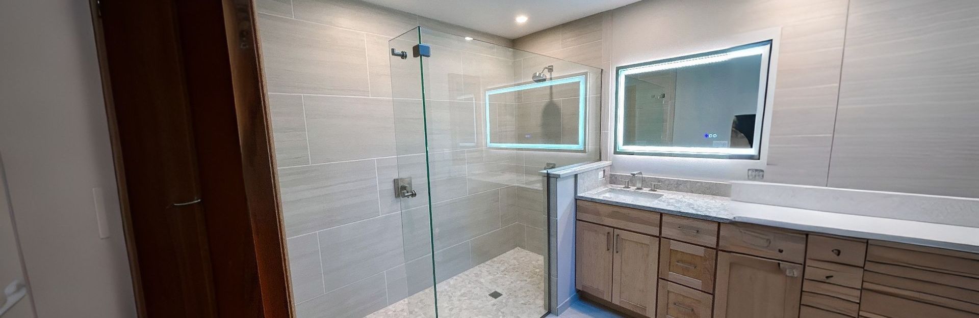 Modern bathroom with a glass shower, two backlit mirrors, and wooden vanity.