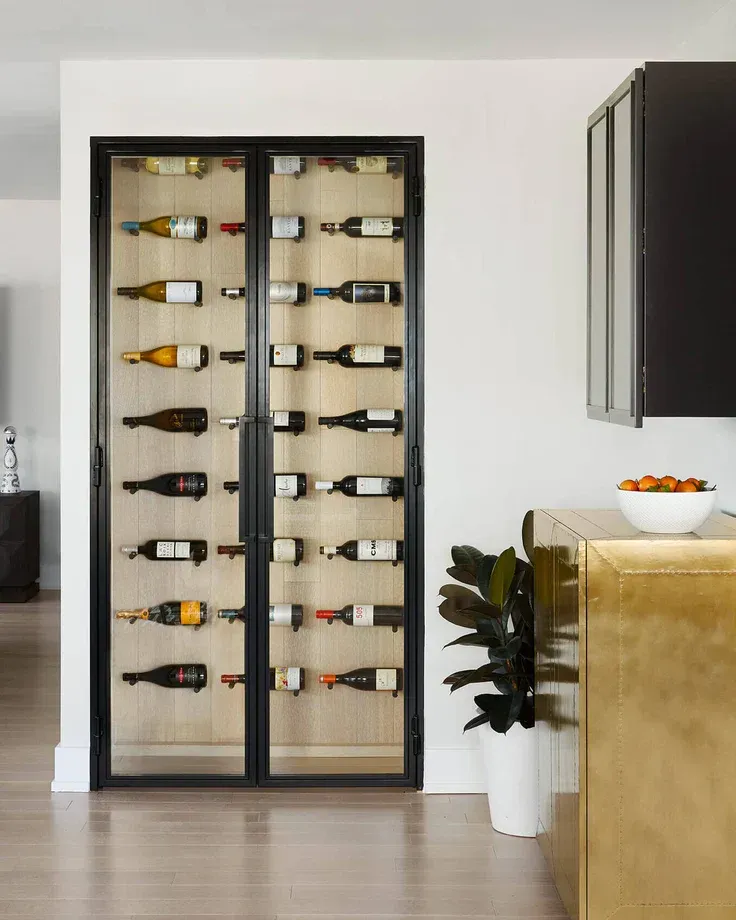 Wine cabinet with black frame and glass doors, filled with bottles, beside a gold cabinet.