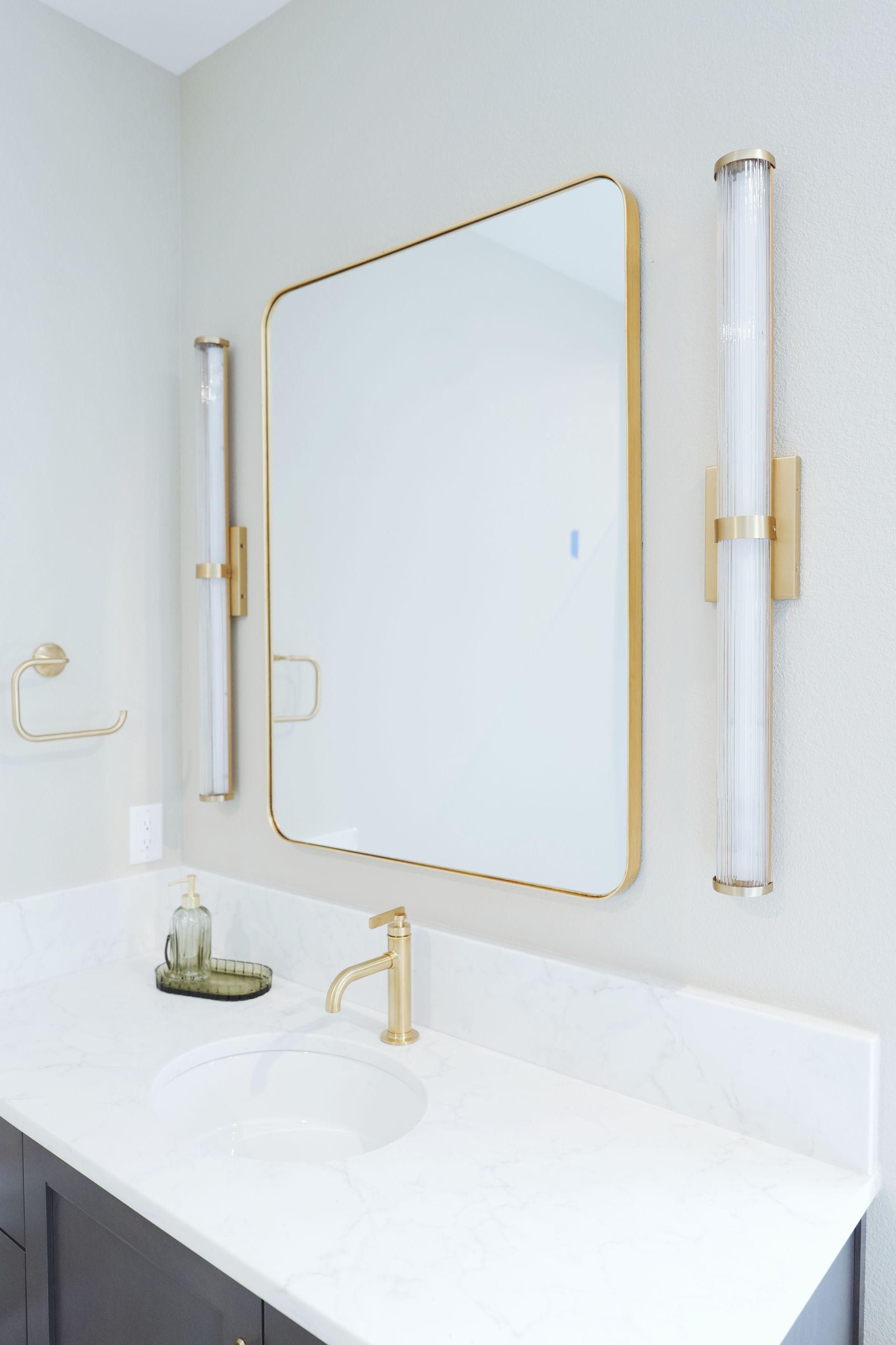 Bathroom with gold accents: mirror, sconces, faucet, and soap dispenser. White countertop and walls.